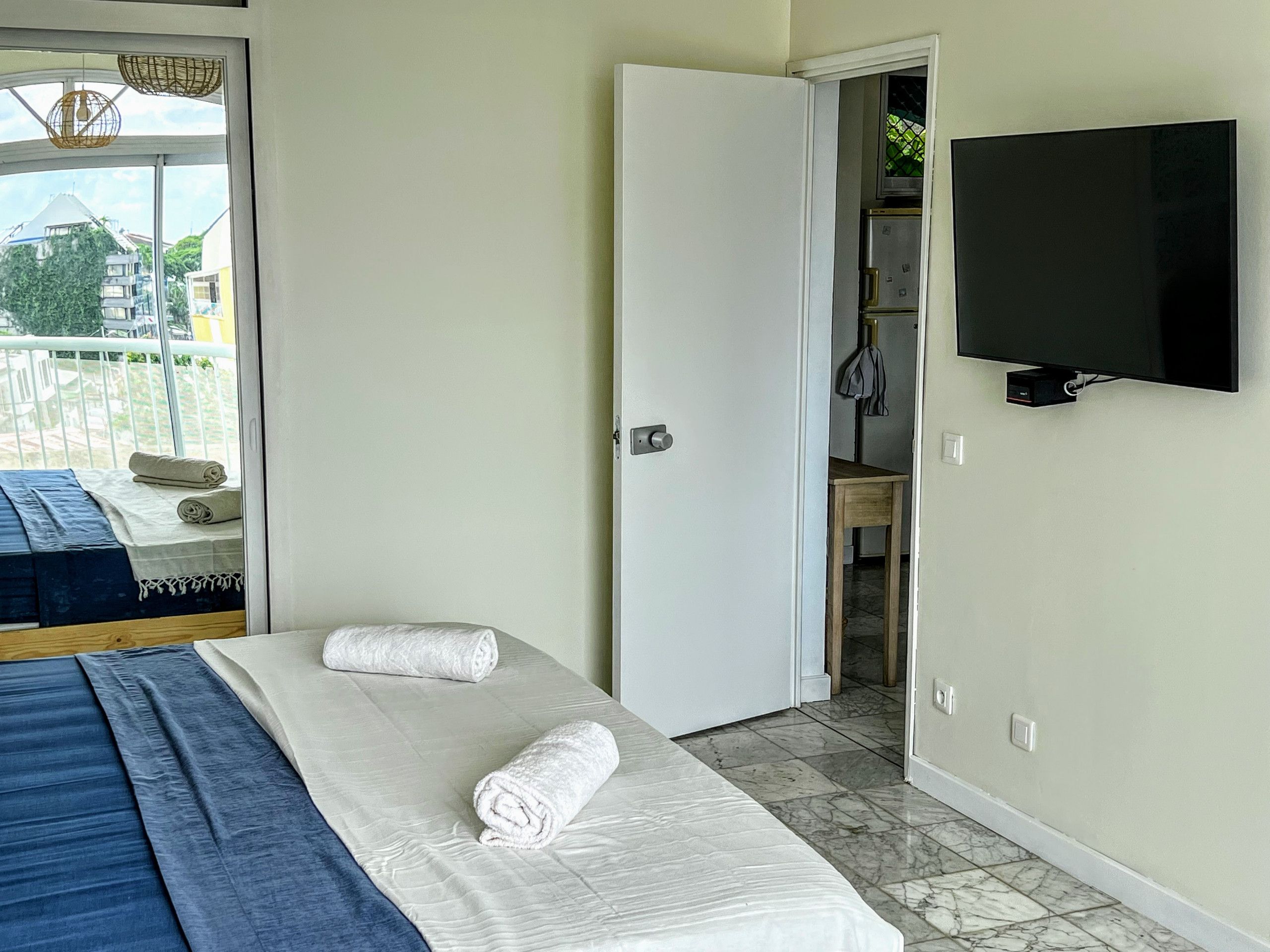 A hotel room with a bed, two rolled towels, a television, and a balcony with a view of buildings.