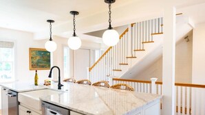 A kitchen with a marble countertop, white cabinetry, and a dining area with wicker chairs. - Luxurious Nantucket Getaway | Heated Pool (Nantucket)