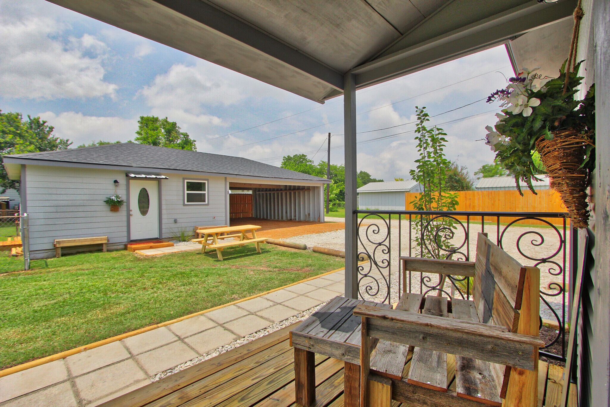 A covered outdoor area with a wooden deck, picnic tables, and a small building in the background.