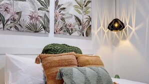 Una cama bien tendida con tres almohadas, una pared con dos estampas botánicas y una lámpara colgante de diseño geométrico.