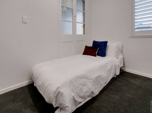 Un dormitorio con cama, paredes blancas, una ventana con persianas y una puerta con vidrio esmerilado.