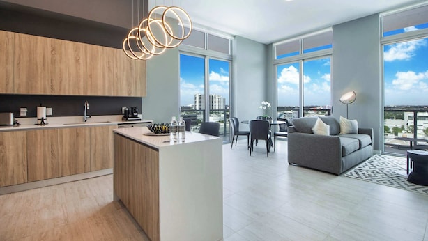 A modern kitchen with a central island, a dining area with a sofa and chairs, and large windows offering a city view. - One Bedroom Condo King Bed With City Views (Miami)