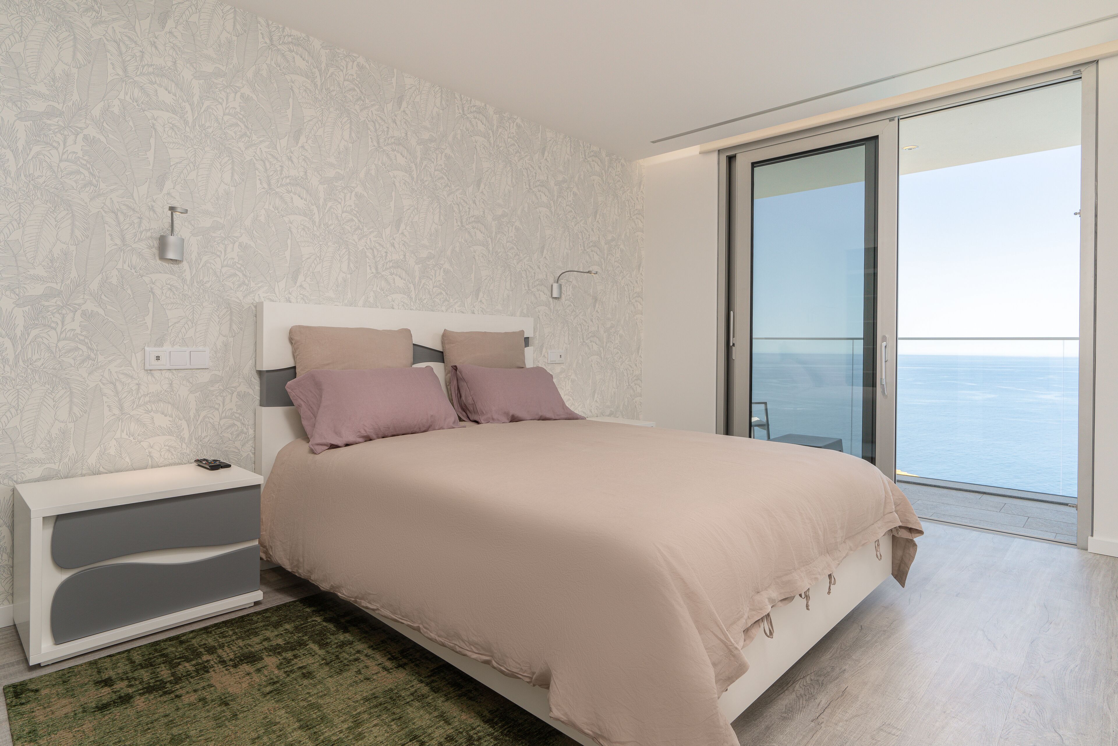 A bedroom with a bed, nightstands, a rug, and a view of the ocean through a glass door.