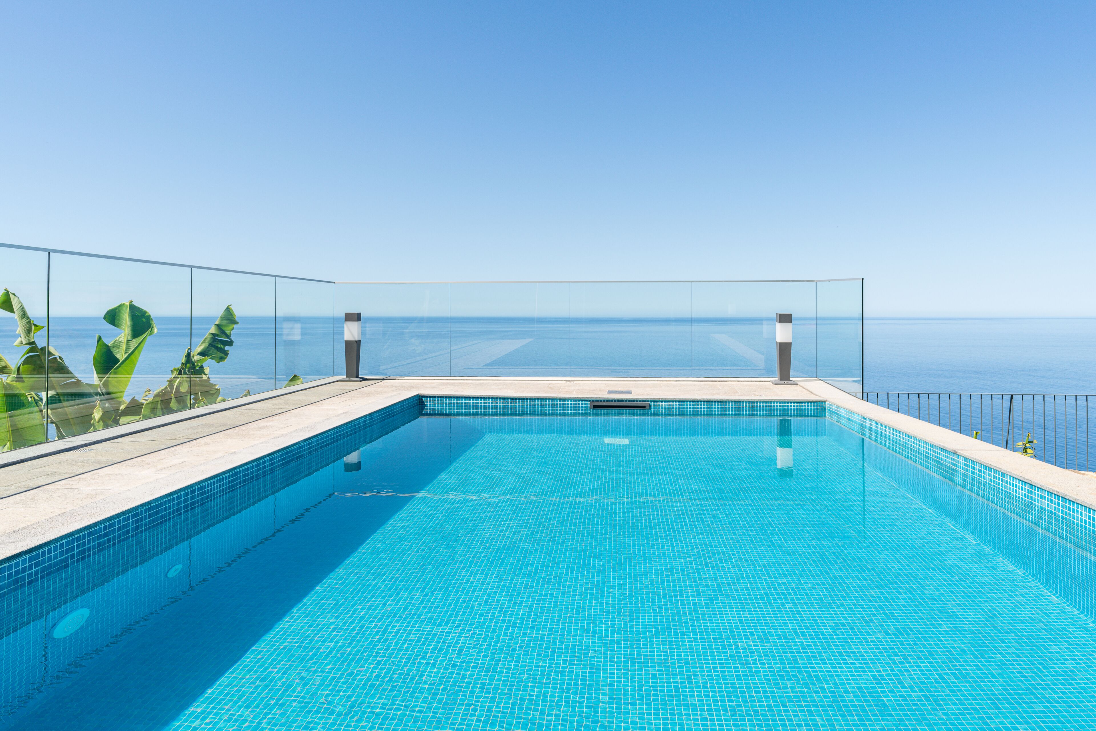 Una piscina infinita azul cristalina con barandilla de vidrio, rodeada de plantas tropicales, con vista a un mar tranquilo.