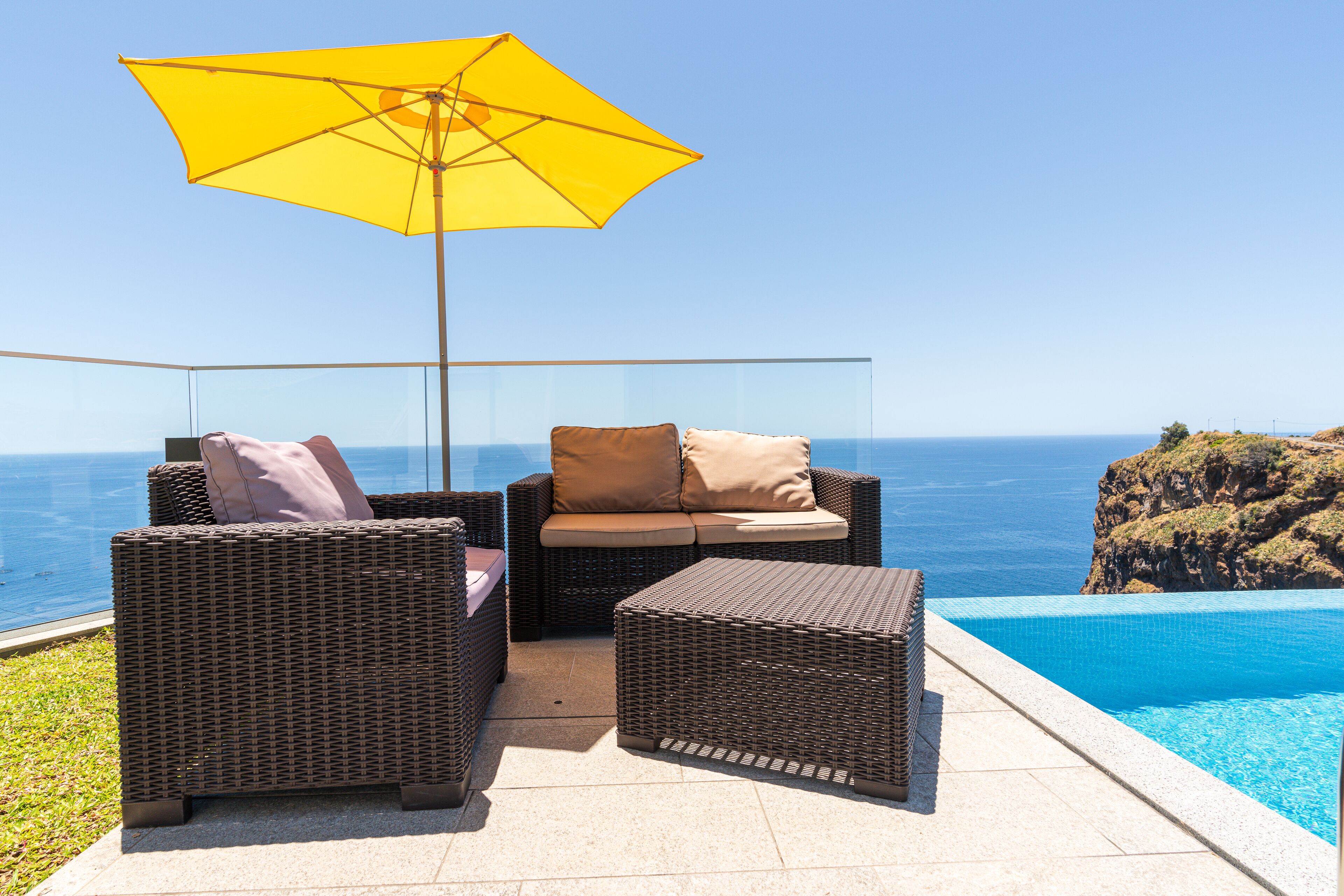 A wicker outdoor seating area with a yellow umbrella, overlooking a clear blue ocean and a rocky cliff.