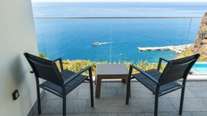 Ein Balkon mit zwei Stühlen und einem kleinen Tisch, der Blick auf die Küste mit Booten und einer felsigen Uferlinie bietet.