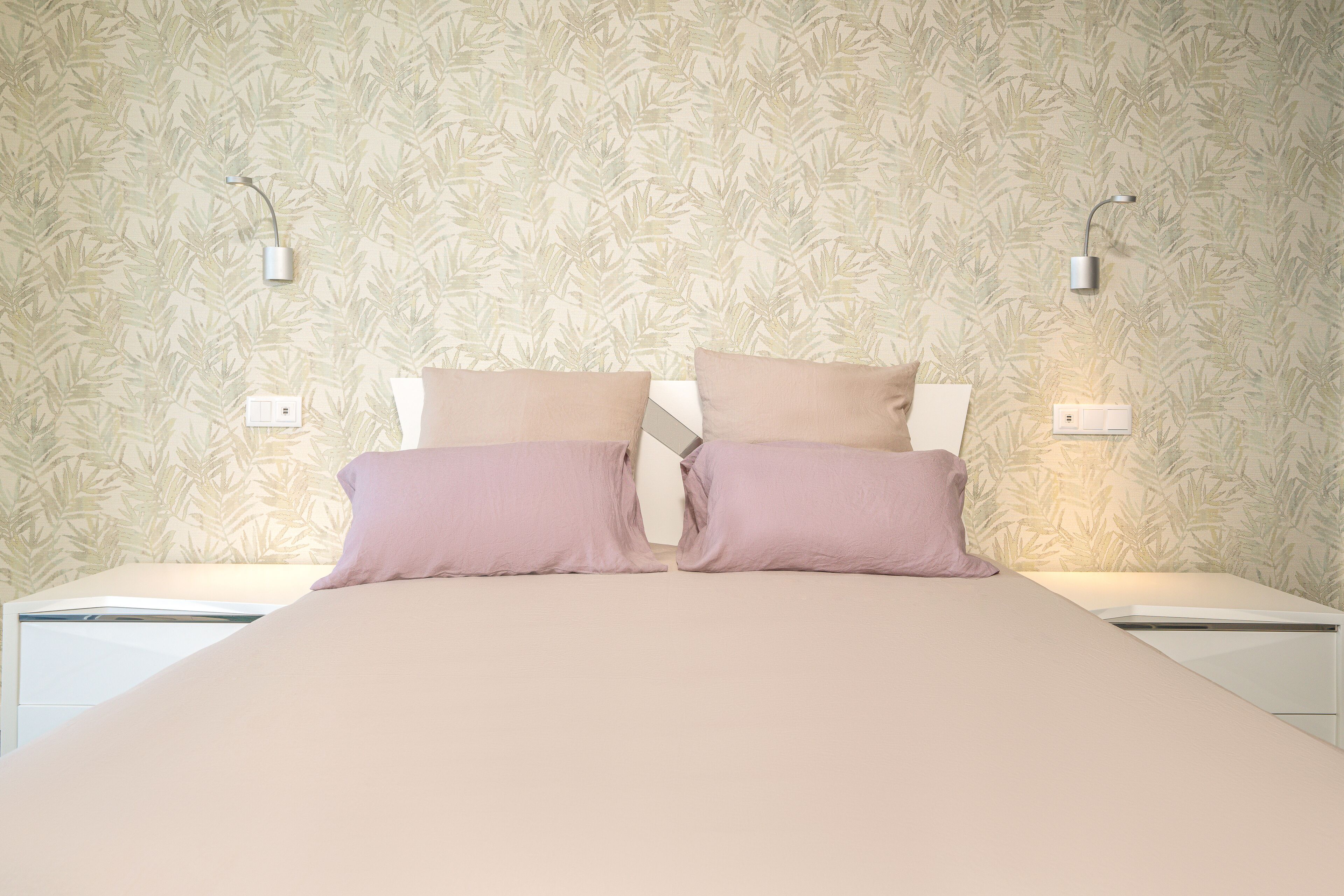 A neatly made bed with two pillows, a wall with a leafy pattern, and two wall-mounted lights.