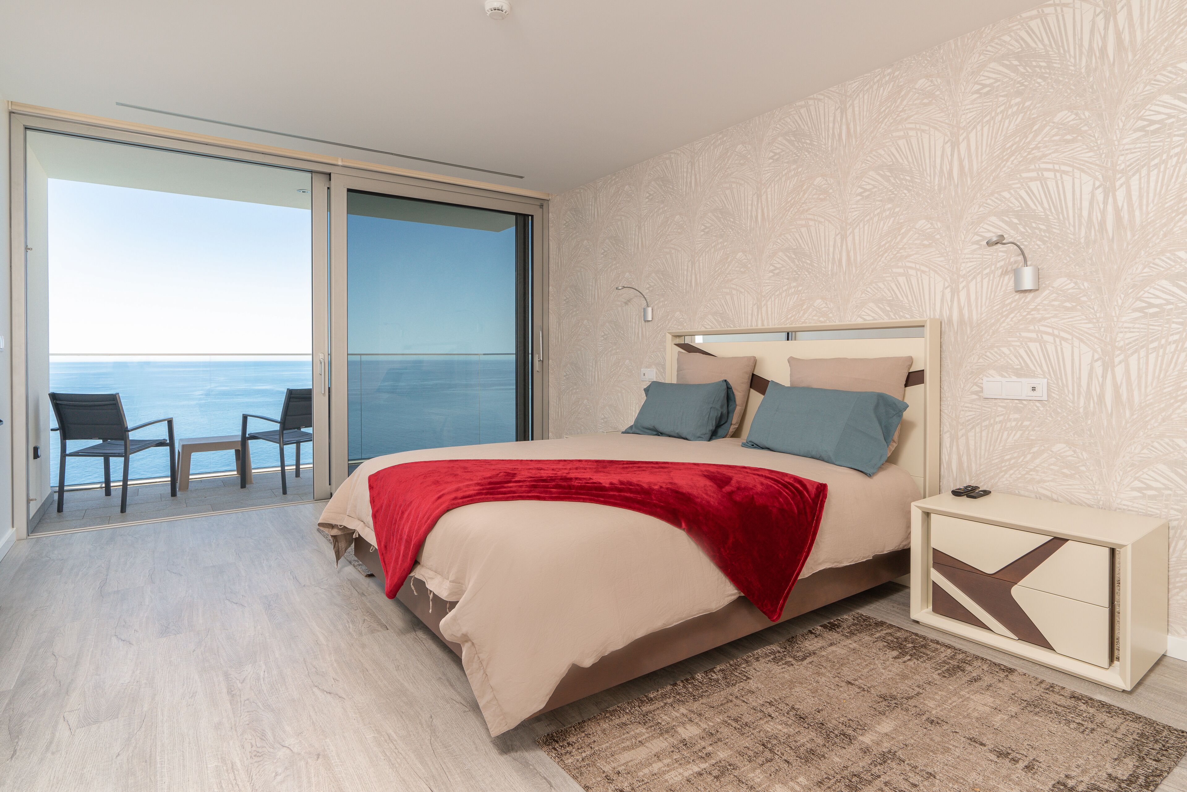 A bedroom with a bed, a nightstand, a chair, and a view of the sea.