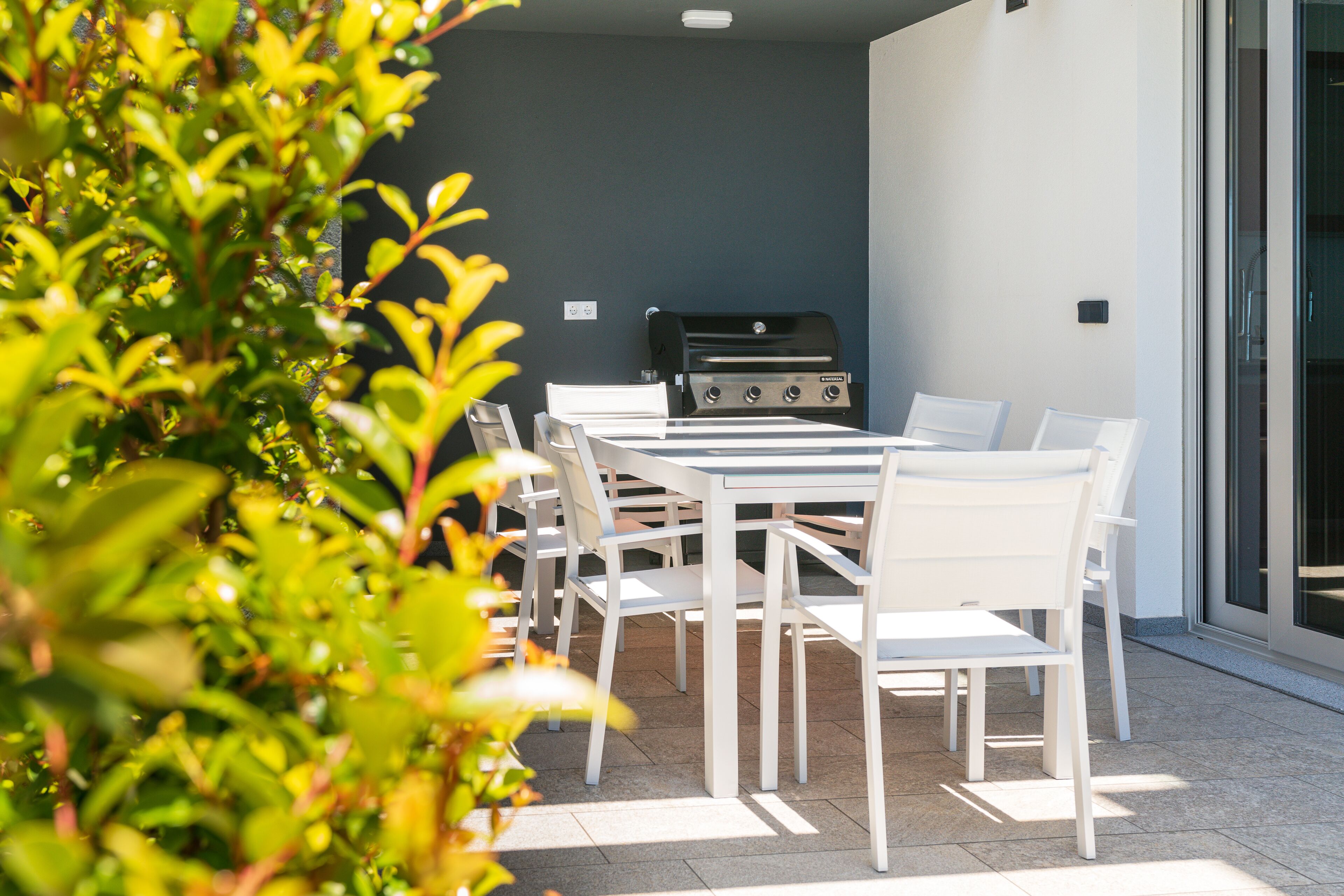 Patio esterno con barbecue, sedie bianche e una porta vetrata che dà su un ambiente interno.