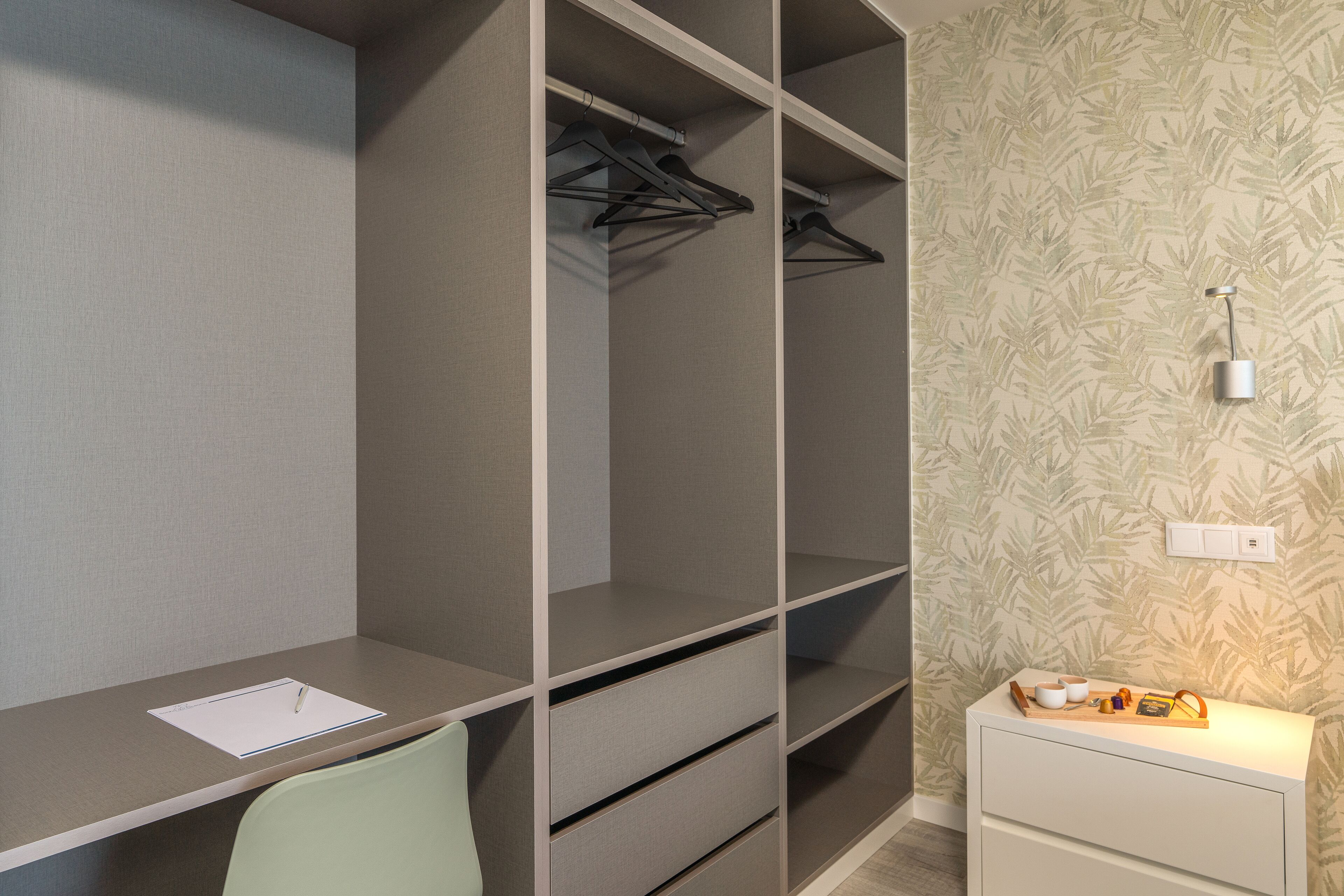 Een kamer met een bureau, een stoel, een kledingkast met meerdere planken en een nachtkastje met een lamp.