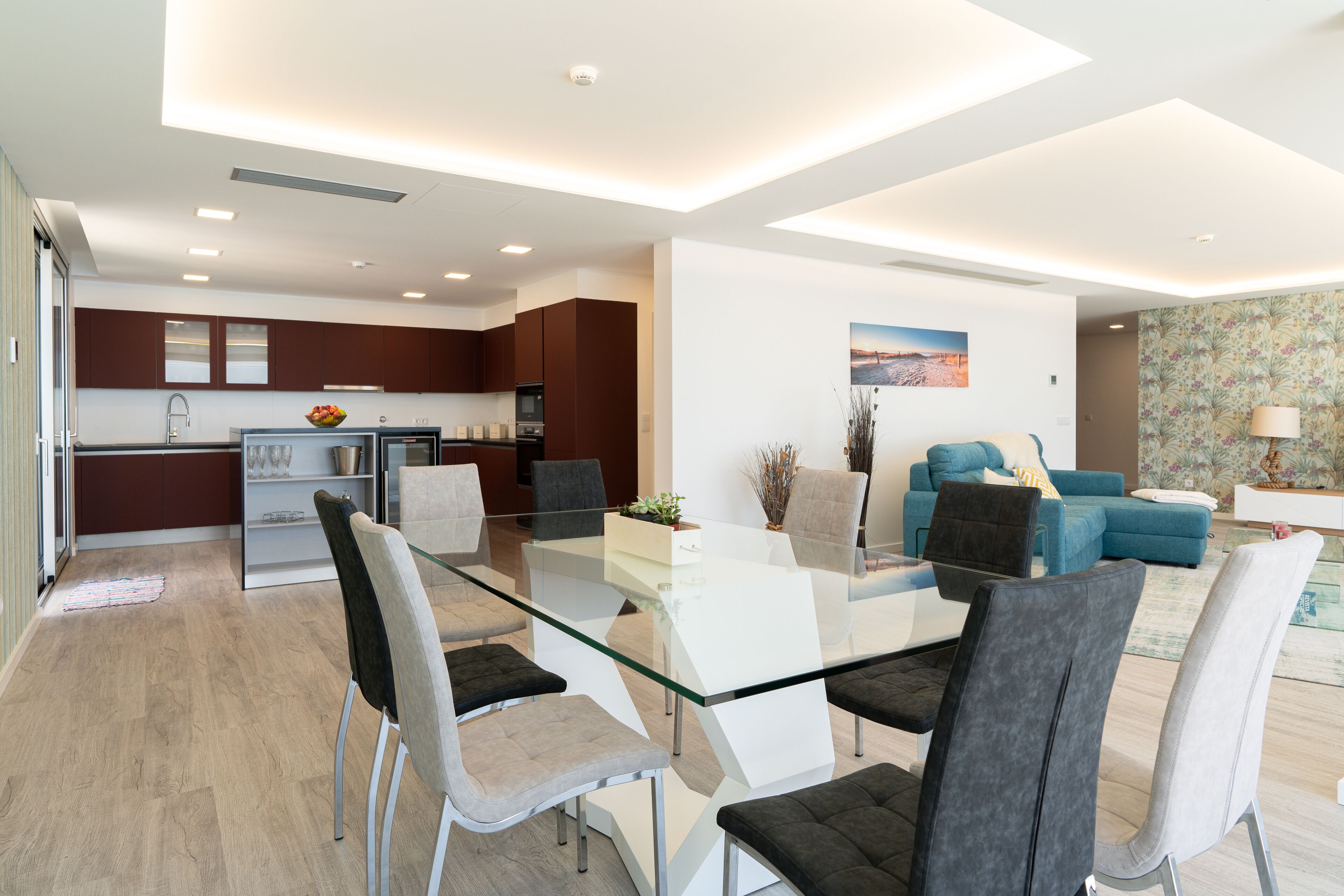 A modern kitchen with dark brown cabinets, a glass dining table, and a seating area with blue sofas and a coffee table.