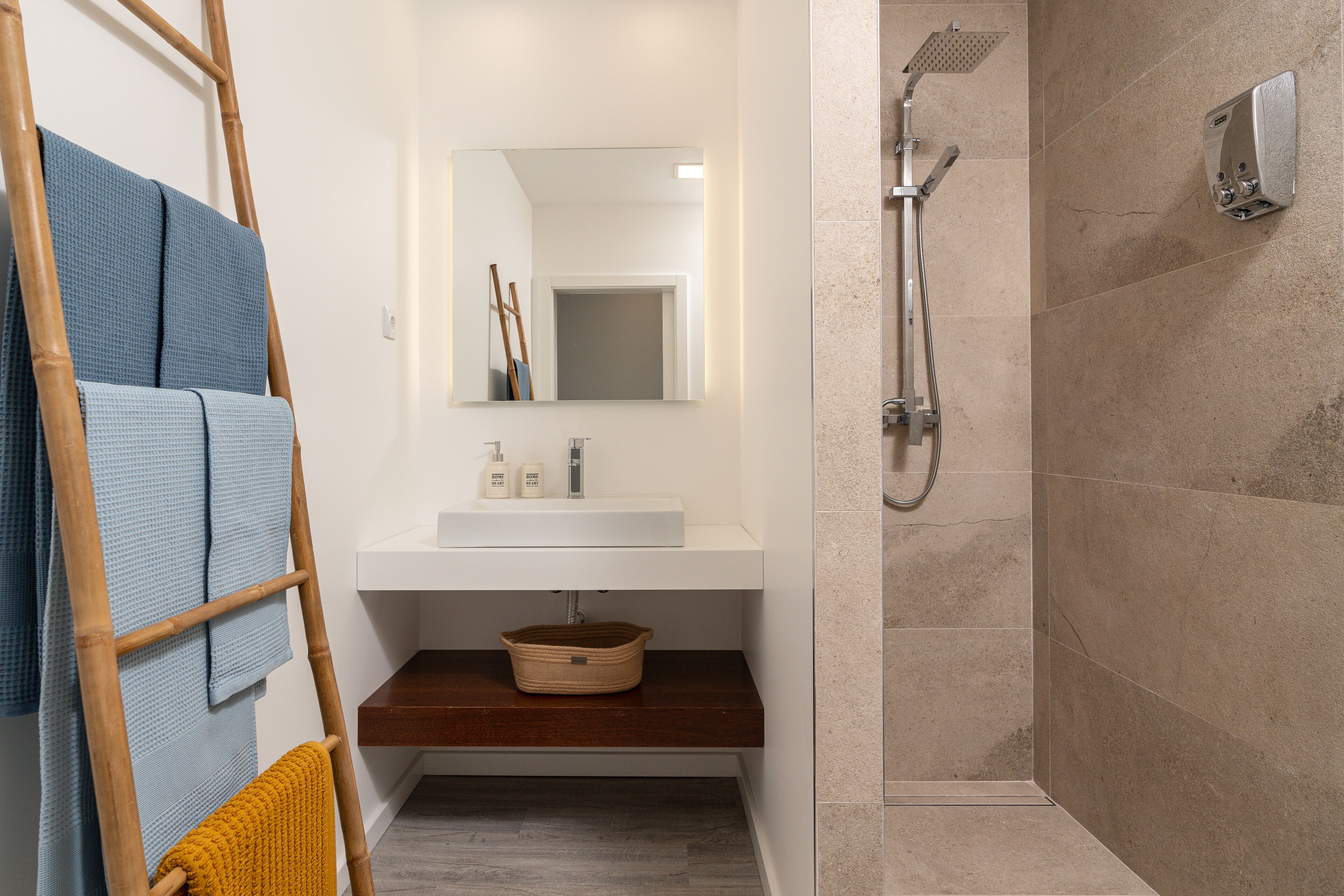 Une salle de bain avec un lavabo blanc, un miroir et une douche. On y trouve une échelle en bois et des serviettes accrochées au mur.