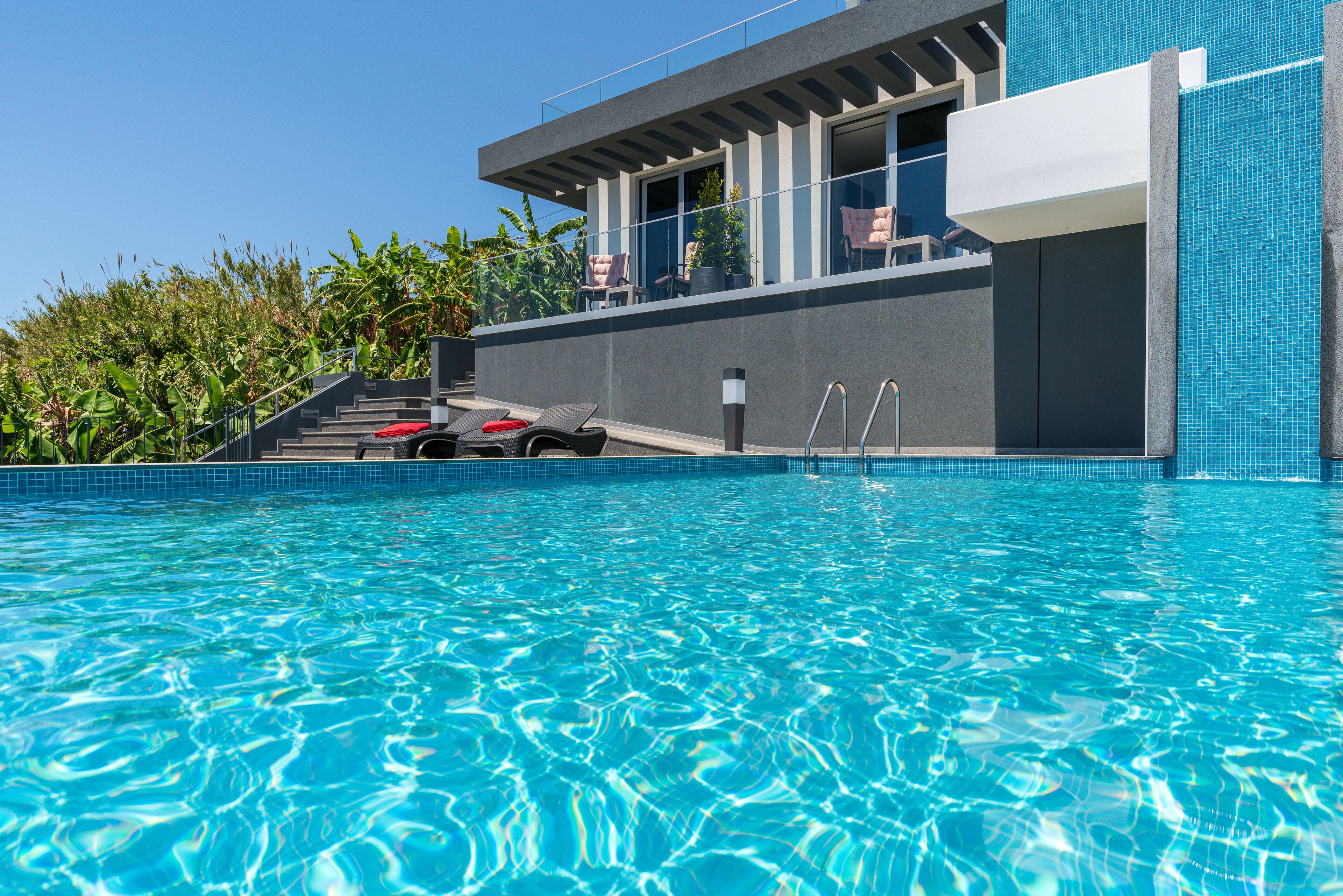 Un área de piscina moderna con agua azul cristalina, una escalera que lleva a un balcón con mobiliario exterior y un edificio con grandes ventanales.
