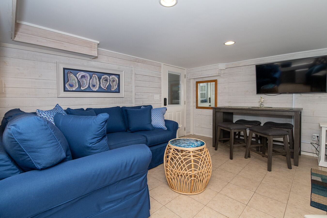 A living room with a blue sofa, a dining table with chairs, a wicker coffee table, and a flat-screen TV mounted on the wall.