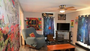 A living room with a grey armchair, a wooden coffee table, a TV on a stand, and a ceiling fan.