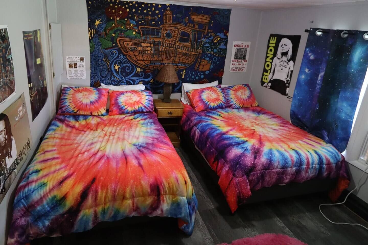 Una habitación con dos camas individuales, cada una con una colcha de estampado psicodélico, un tapiz en la pared y carteles.