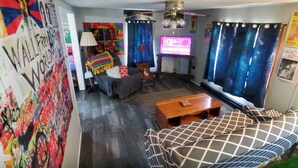 A room with a bed, a sofa, a TV, and a wall with colorful artwork.