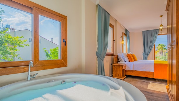 A bedroom with a large bathtub, a bed with orange pillows, and a view of the outside. - Myra Villa 1 (Fethiye)