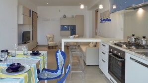Et moderne køkken med en central ø, indbygget ovn og stålstål-hvidevarer. Der er et spiseområde med et bord dækket til fire personer, gennemsigtige barstole og et fjernsyn på et skab.
