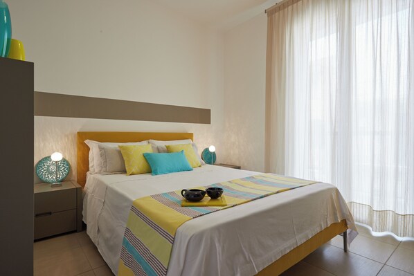Una cama bien tendida con una colcha a rayas, una mesita de noche con lámpara y una ventana con cortinas de gasa.