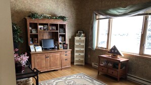 A room with a wooden cabinet with shelves, a television, a small table with a model, a patterned rug, and a window with curtains. - Spacious home, large deck with beautiful views in the heart of the Black Hills  (Rapid City)