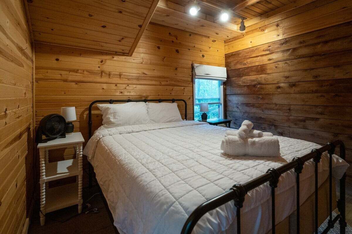 Ein Zimmer in einer Holzhütte mit einem Bett, einem Nachttisch, einem Fenster und einem Ventilator.