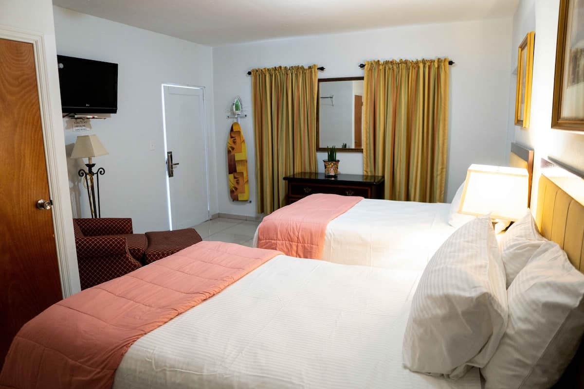 A hotel room with two beds, a television on the wall, a mirror, and a window with curtains.
