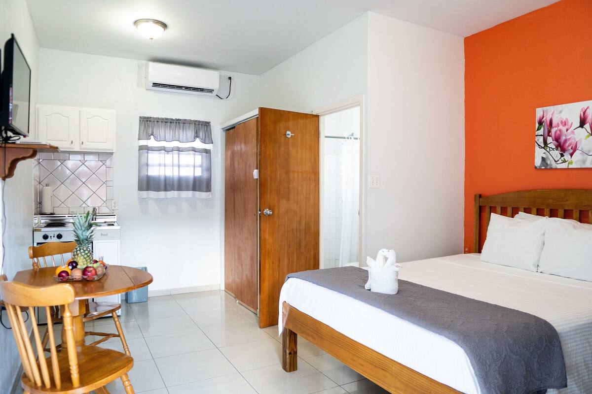 A bedroom with a wooden bed, a small dining table with fruit, a kitchen area with a stove, and a white wall with a floral painting.
