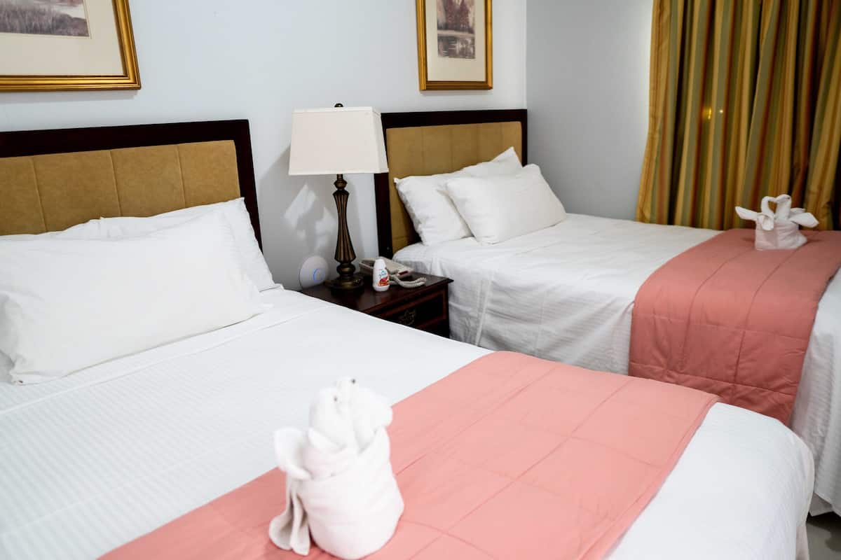 A hotel room with two beds, each with white bedding and a pink accent. There are framed pictures on the wall and a lamp on the nightstand.