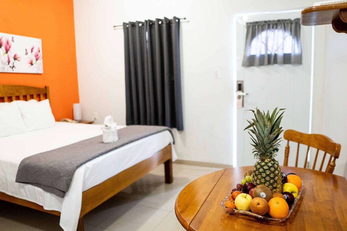 A hotel room with a bed, a wooden table with a fruit basket, and a window with curtains.