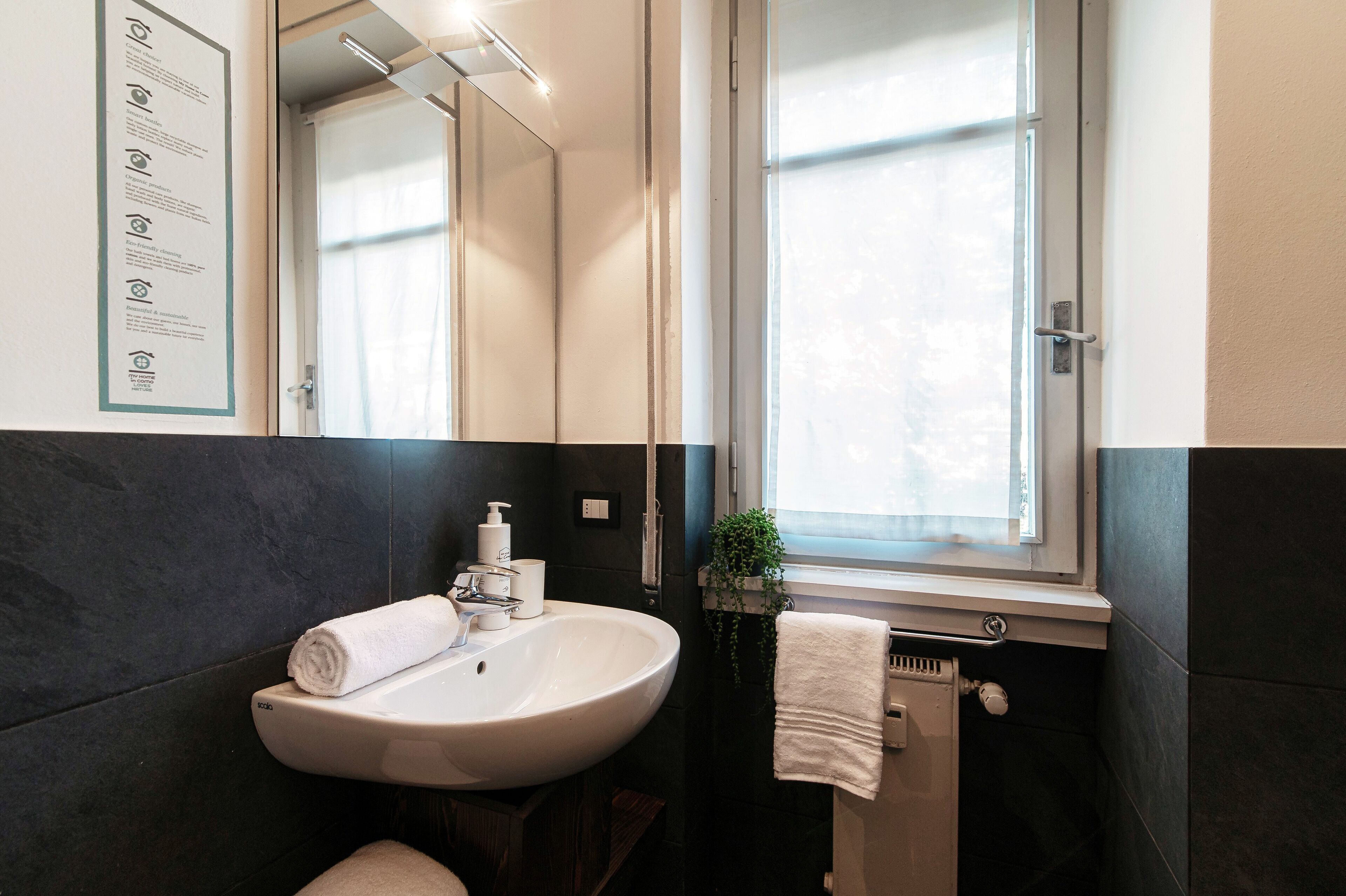 Ein Badezimmer mit einem weißen Waschbecken, dunklen Fliesenwänden, einem Fenster und einem Spiegel.