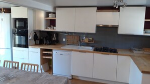 A compact kitchen with white cabinets, a built-in oven, microwave, and dishwasher. There is a wooden dining table with chairs, and a small window providing natural light. - Gîte Pour 8-10 Personnes Avec vue Imprenable ! (Gérardmer)