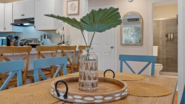 Un comedor con una mesa de madera, sillas azules, un florero de vidrio con una planta y una cocina al fondo.