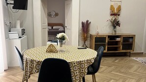 A dining area with a round table, black chairs, and a patterned tablecloth. A television is mounted on the wall, and a wooden cabinet with a macrame decoration is visible.