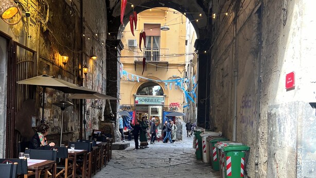 A narrow alley with stone pavement, wooden tables, and chairs, leading to a building with a yellow facade and an arched entrance.