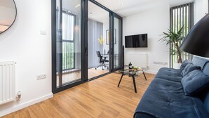 Un salón moderno con un sofá azul, un ventanal de vidrio que da a un balcón, un televisor montado y una mesita con un jarrón.