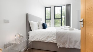 Un dormitorio moderno con una cama, una mesita de noche despejada, una ventana con cortinas y una puerta de madera.