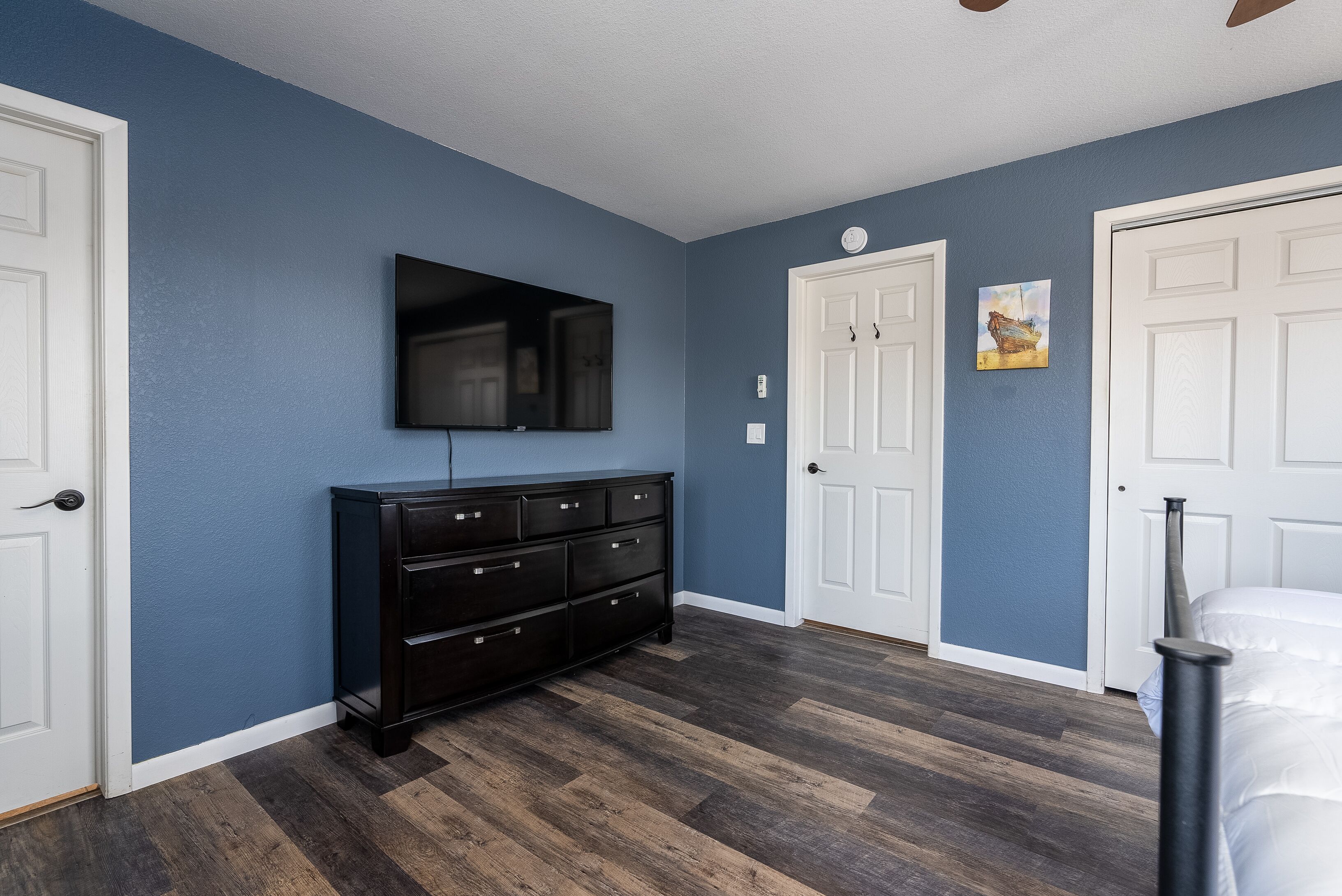 A room with blue walls, a wooden floor, a black dresser, a flat-screen TV, and white doors.