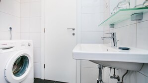 Moderni kylpyhuone, jossa on Bosch-merkkinen pesukone, valkoinen pesuallas ja lasihylly pesualtaan yläpuolella.
