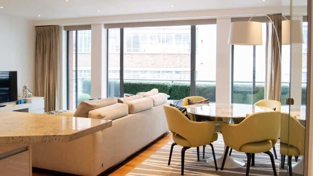 Uma sala de estar moderna com sofá, mesa de jantar com cadeiras, janelas amplas e televisão de tela plana.
