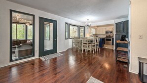 A spacious open-plan living area with a dining table and chairs, a kitchen area with cabinets and appliances, and a large window providing natural light. - Mountain Bikers Retreat (Bella Vista)
