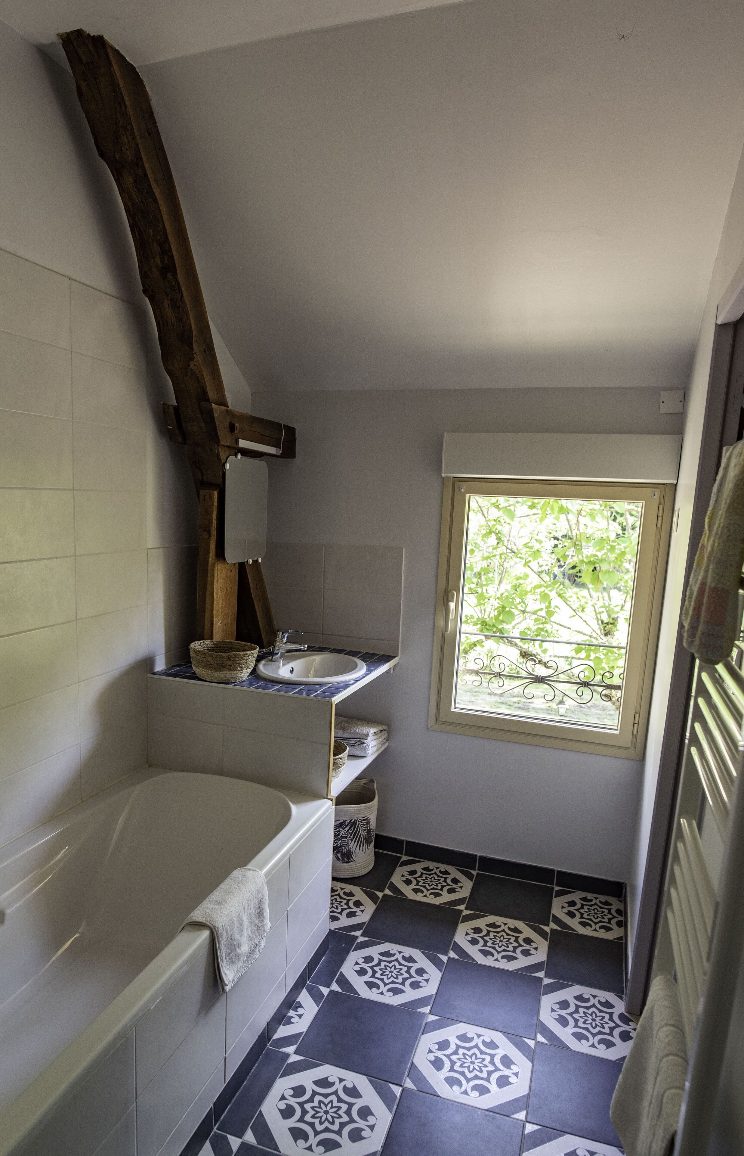 Une salle de bain avec une baignoire, une fenêtre donnant sur un paysage verdoyant et des carreaux de sol à motifs.