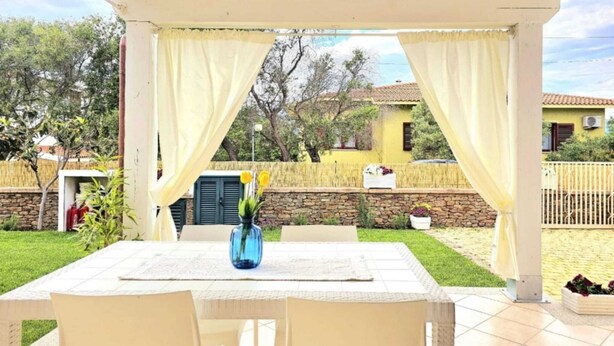 A covered patio with a table and chairs, a vase with flowers, and a view of a garden and buildings in the background. - Casa Maria (Tanaunella)