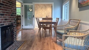 Une pièce spacieuse avec un plafond en bois, une cheminée en briques, une table à manger avec des chaises, des meubles en osier et une grande fenêtre.