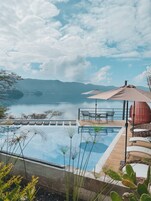 Una piscina infinita con vistas a un lago y montañas, mobiliario exterior y una terraza de madera.