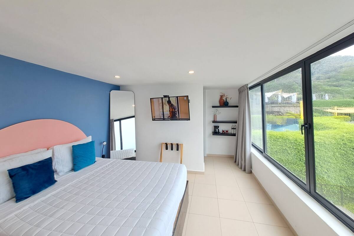 A modern bedroom with a large bed, a TV, and a view of a green landscape through a large window.