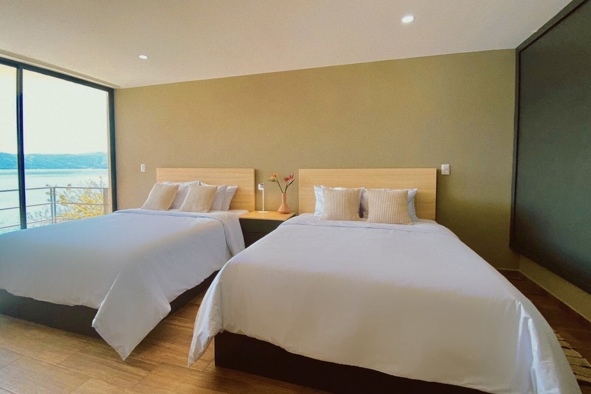 A hotel room with two beds, a nightstand, a vase with flowers, and a view of the outdoors.