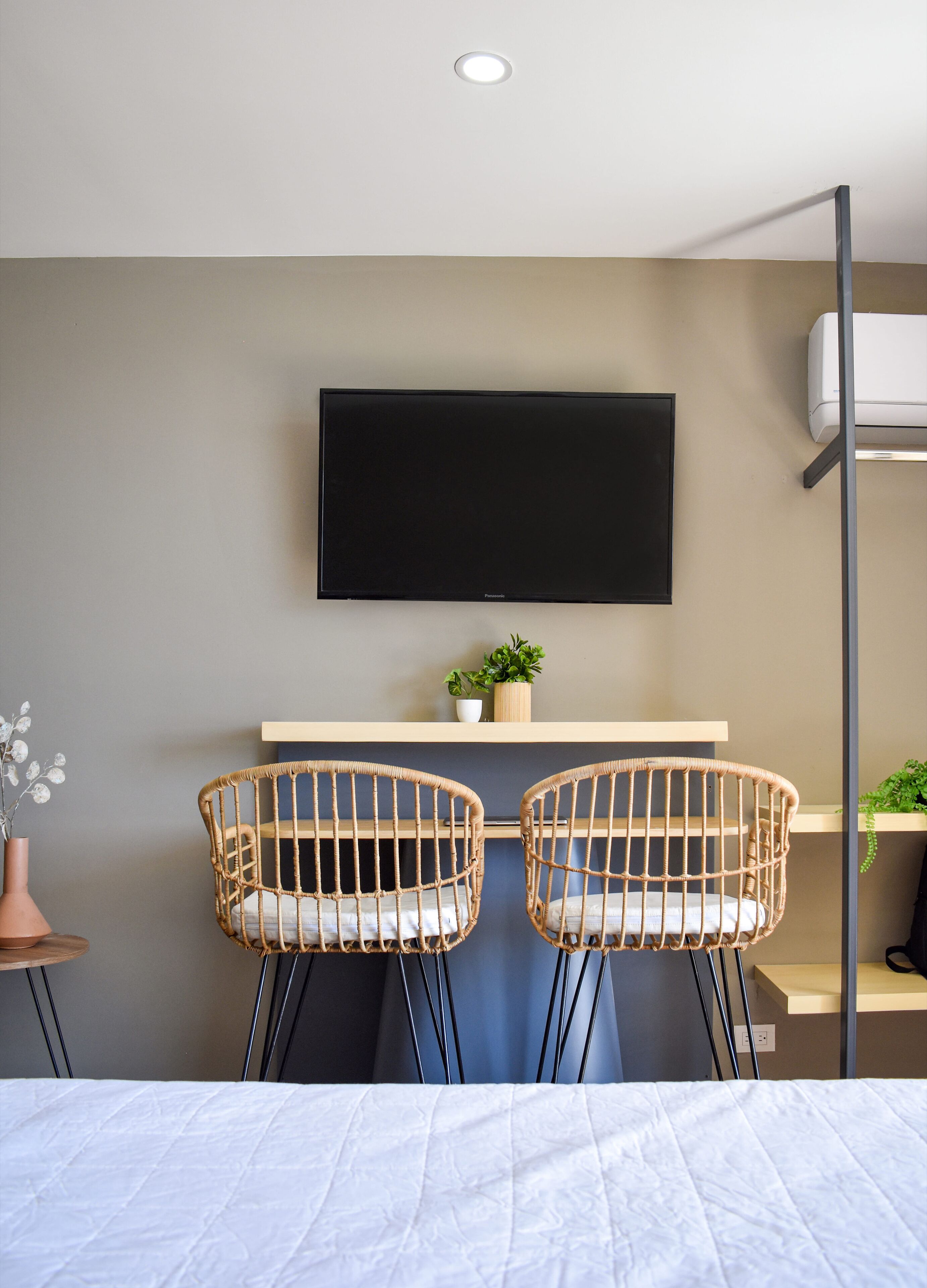 Une chambre d’hôtel moderne équipée d’un lit, de deux fauteuils en osier, d’un téléviseur à écran plat, d’une petite plante et d’un système de climatisation intégré.