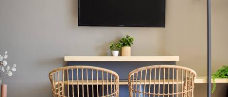 Une chambre d’hôtel moderne équipée d’un lit, de deux fauteuils en osier, d’un téléviseur à écran plat, d’une petite plante et d’un système de climatisation intégré.
