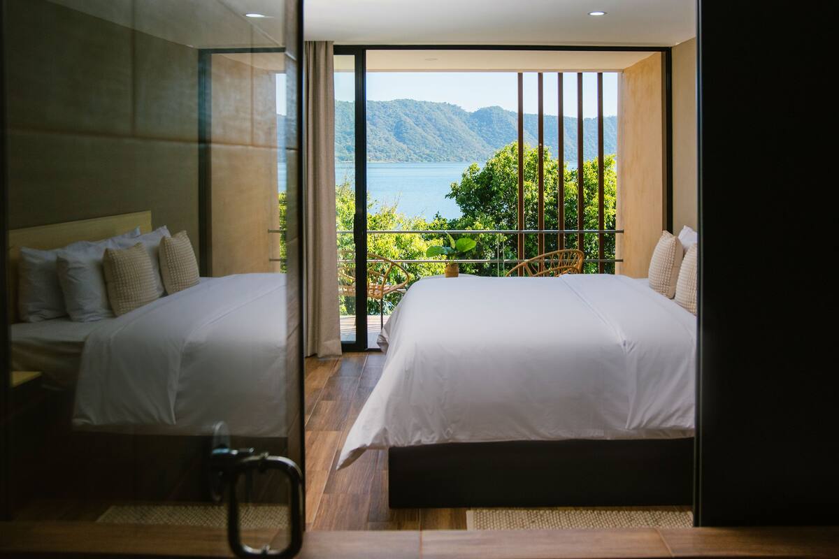 A modern hotel room with two beds, a balcony with a view of a lake and mountains, and a sliding glass door.